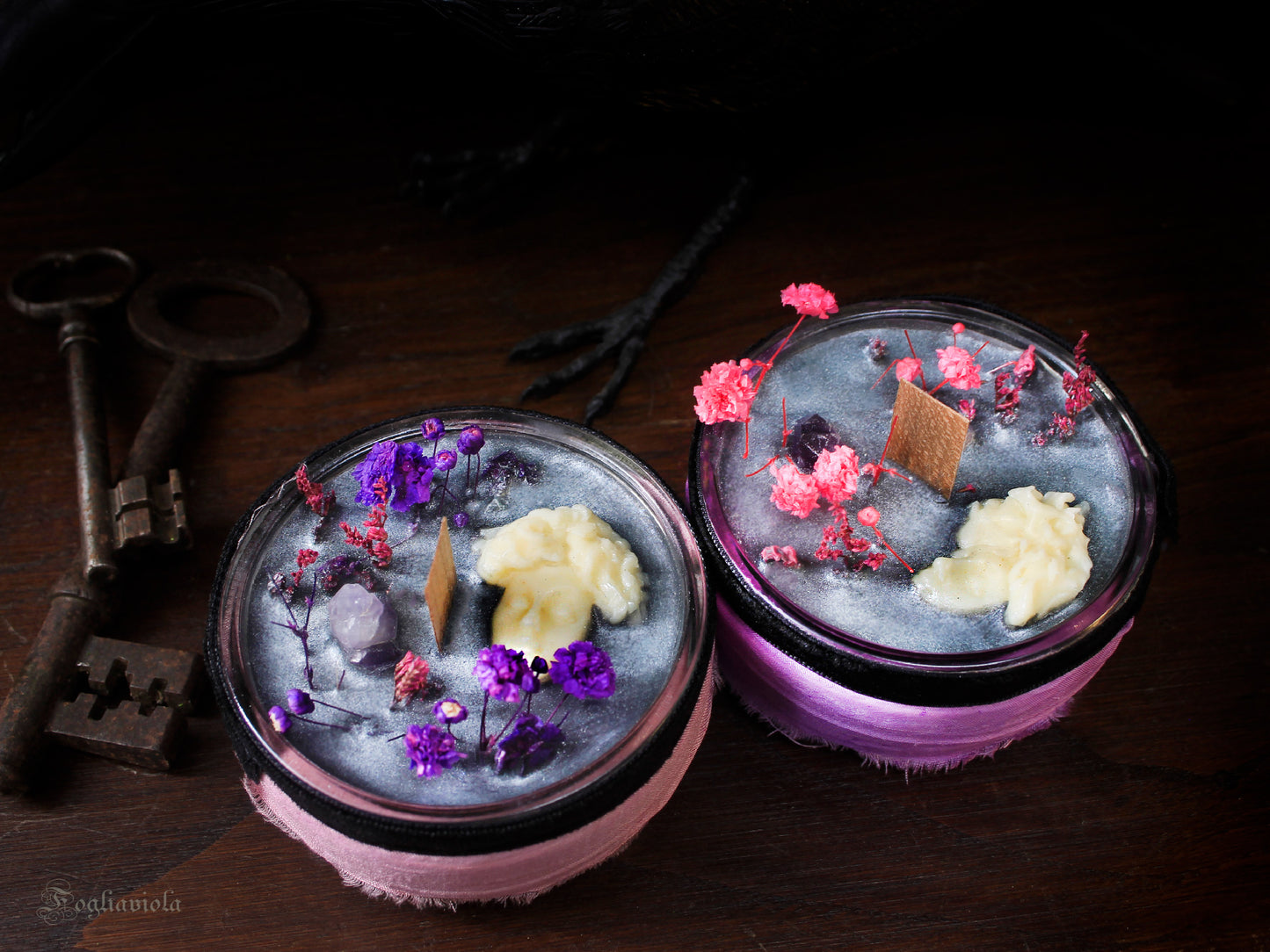 Candela votiva Ecate in cera argento-cenere con testa di dea scolpita, ametista grezza, fiori viola e rosa essiccati e stoppino di legno, accanto a chiavi antiche in ferro.